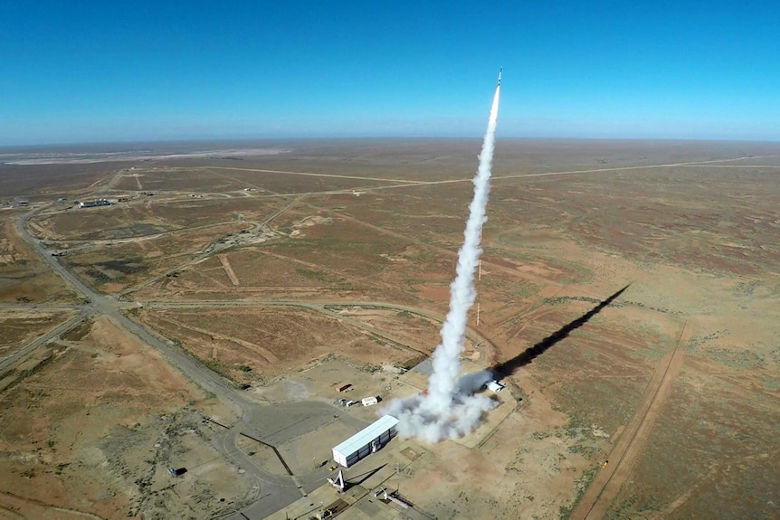 An atmospheric photo shows a rocket below launched with a vertical column of smoke behind it falling toward brown landscape.