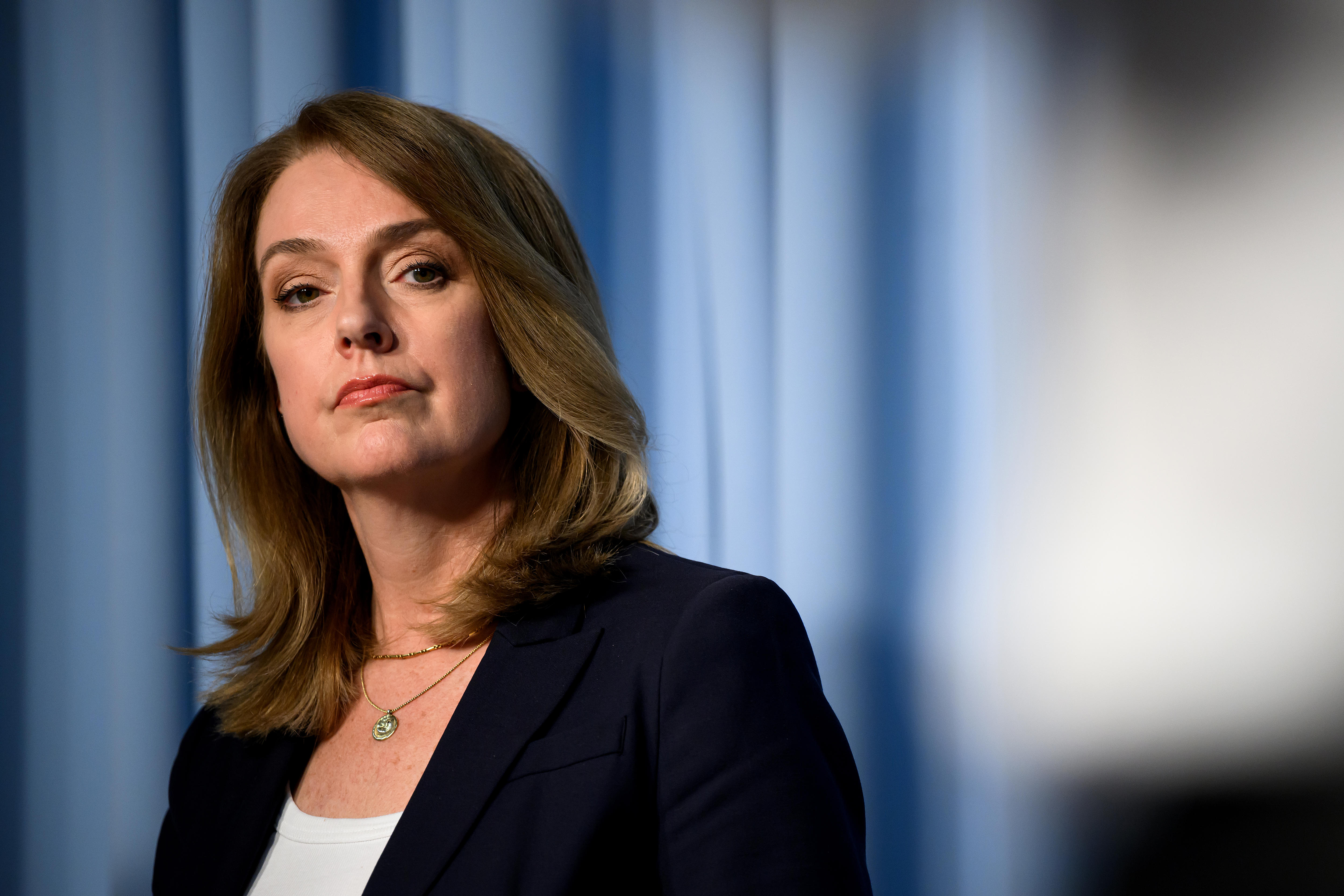 Uma mulher com cabelo castanho de comprimento médio e uma jaqueta preta olha com confiança para a câmera enquanto outra pessoa fala.