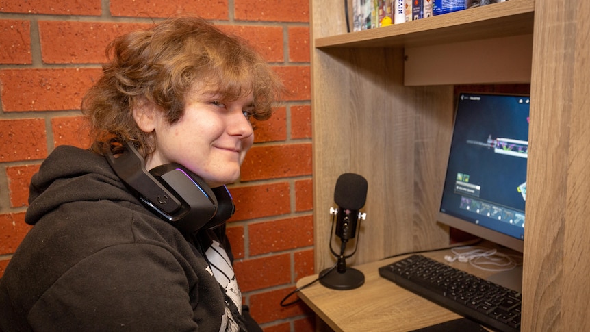 young person with fair skin smiles at the camera sitting at desk next to a computer