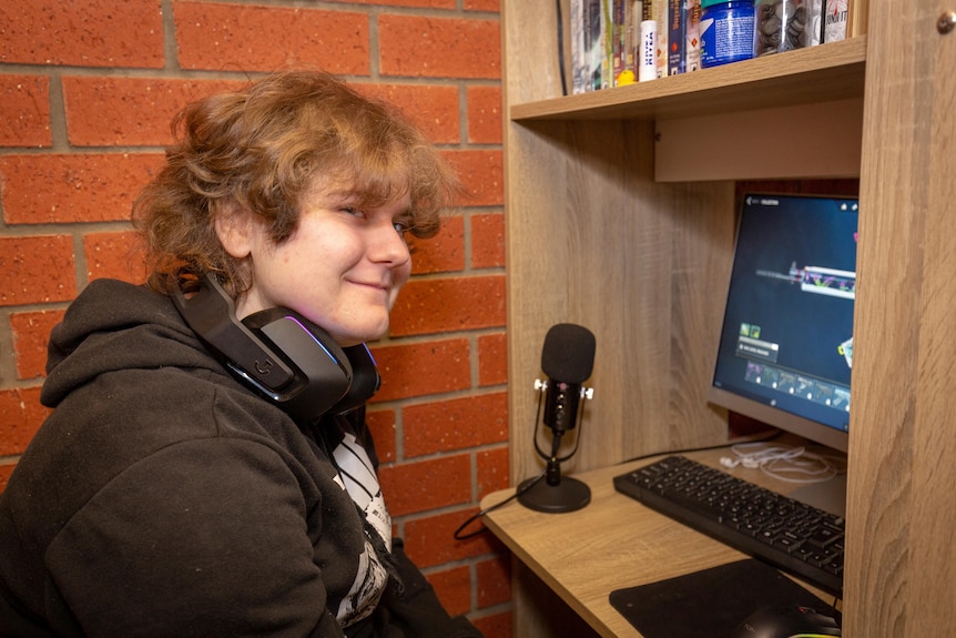 A young person with fair skin smiles at the camera sitting at desk next to a computer