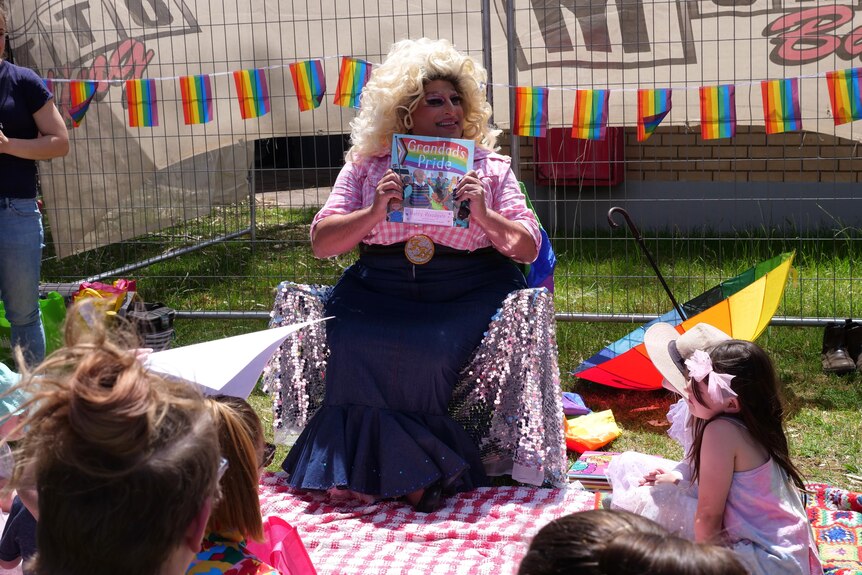 A drag queen reads a book to some children outside a library.