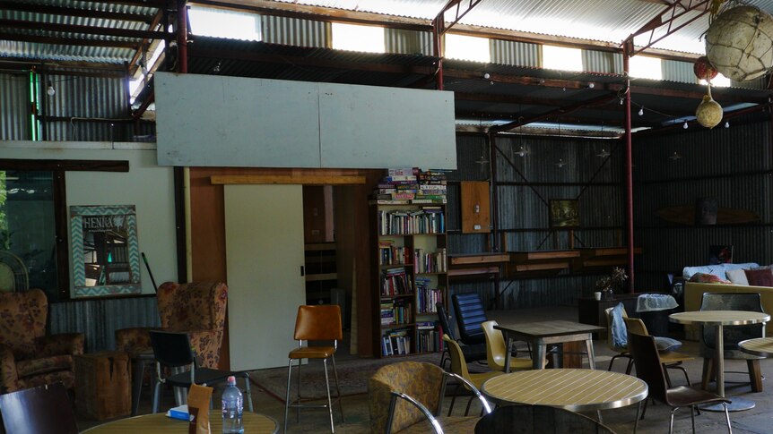 A picture inside a rustic shed-like building. It's filled with tables and chairs and a bookcase.
