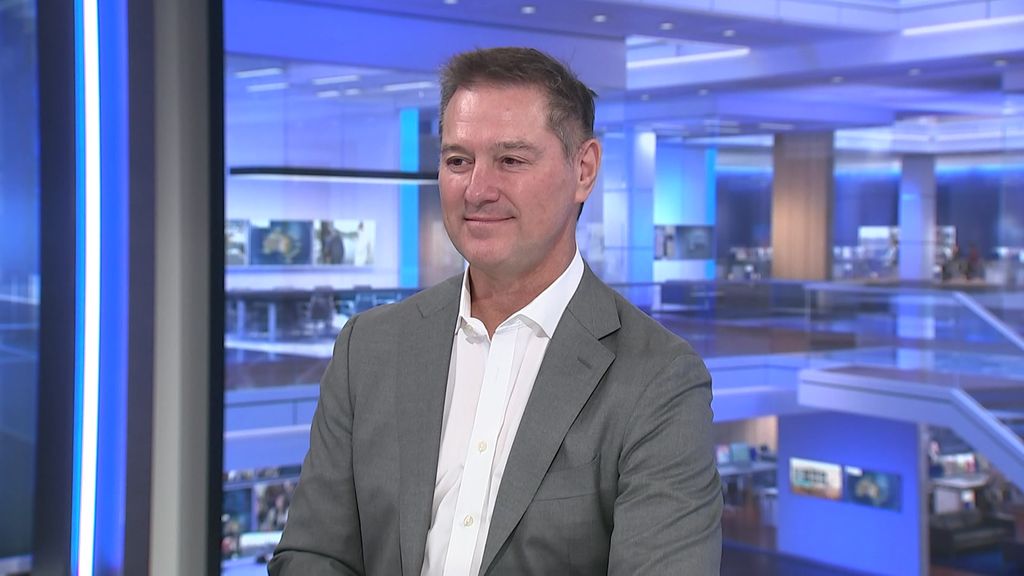 A man with grey hair smilin while seated in a television studio during a news interview.