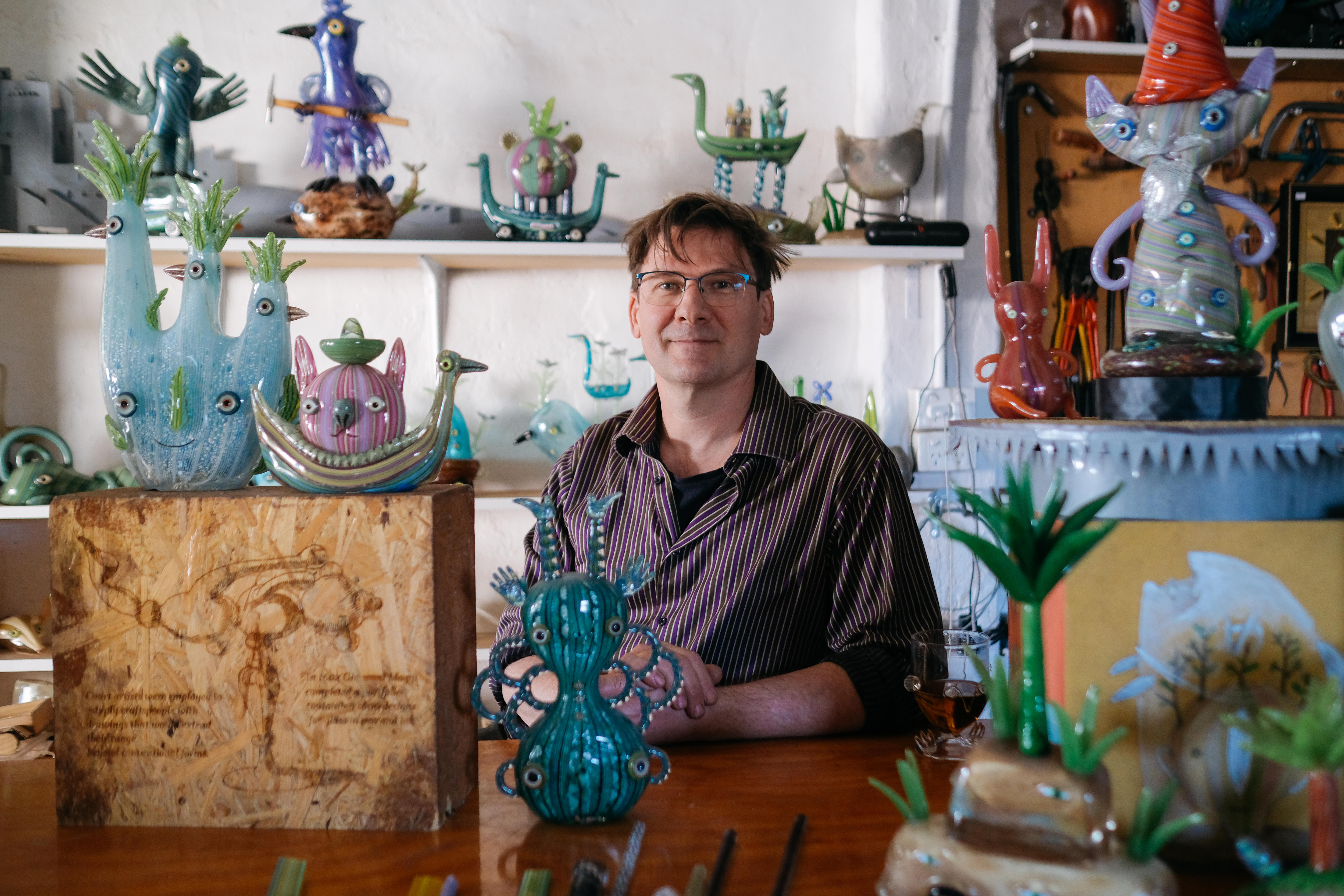 Artist Tom Moore in his studio, surrounded by his colourful glass creations. He is middle-aged with brown hair and glasses 