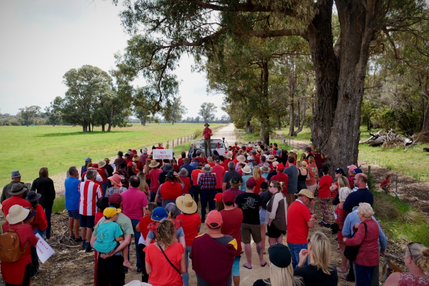 A hundred people gathering next to a paddock and listing to a man speak who is standing on a ute.