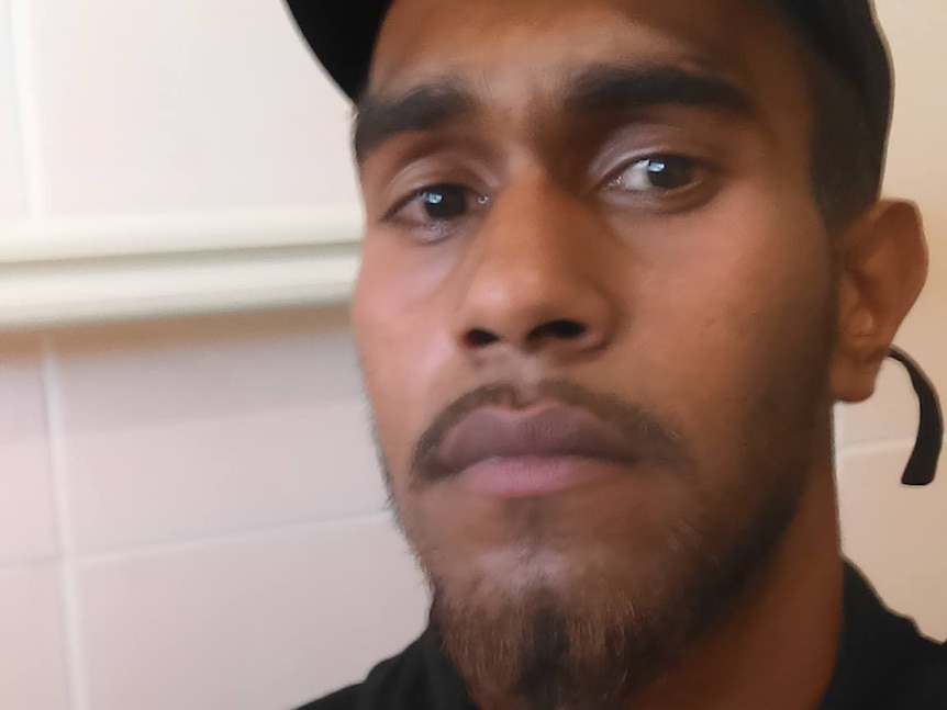 Jaycob wears a black cap and black top. He is standing in front of a tiled wall.