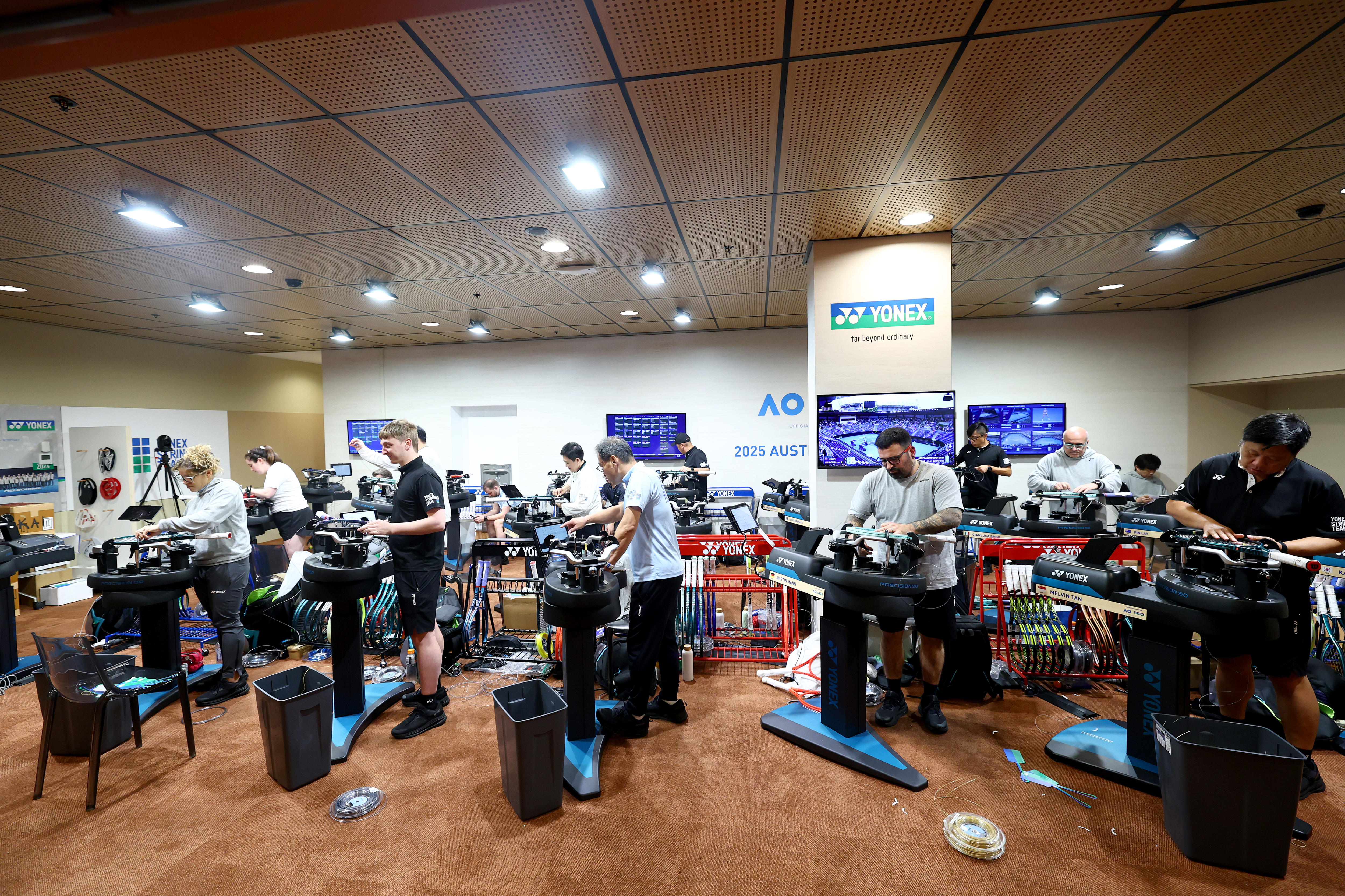 Várias pessoas trabalham em máquinas de encordoamento de raquetes.