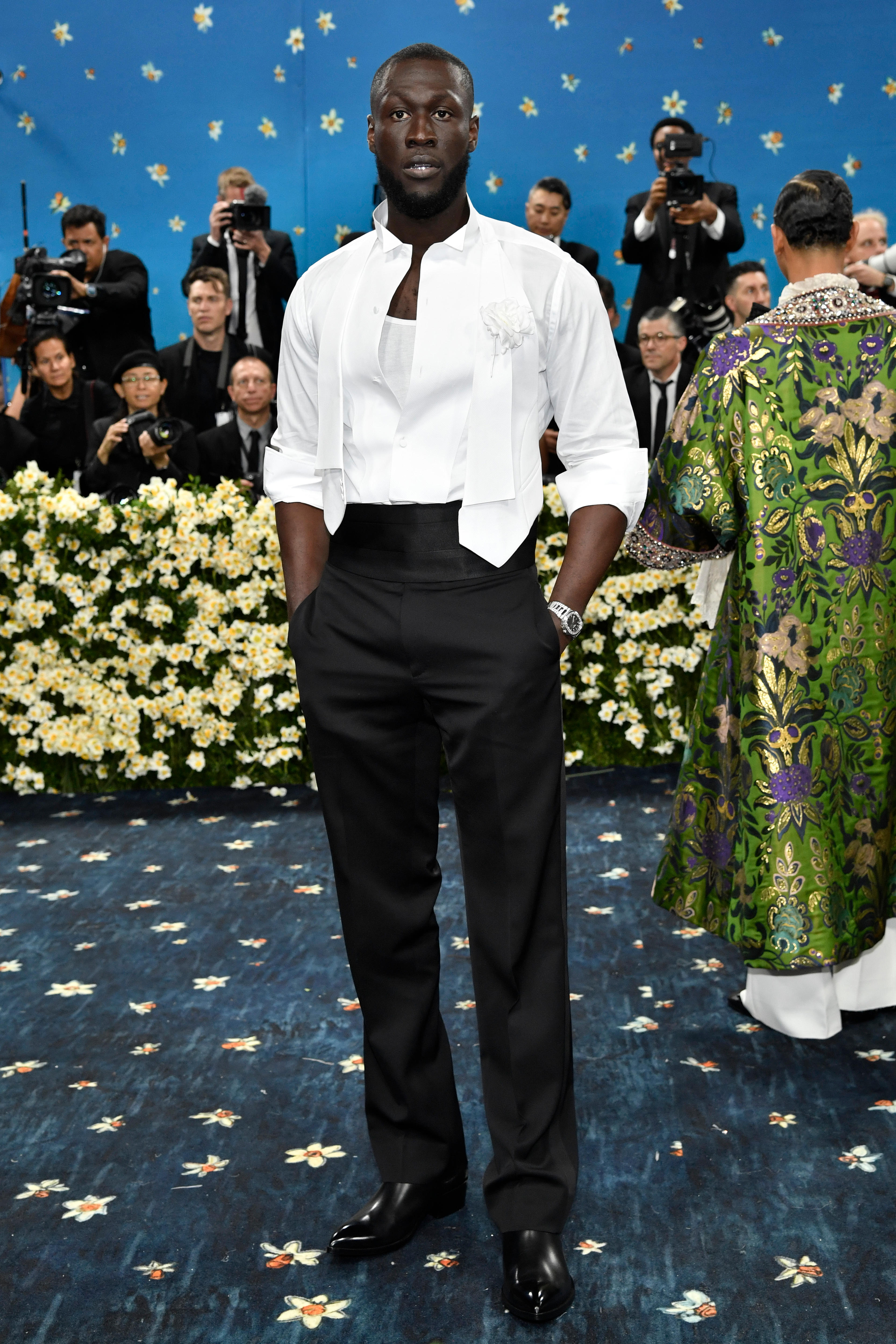 Stormzy wearing black trousers and an open white button up shirt top with pushed up sleeves