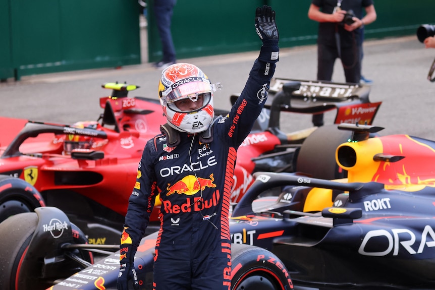 Racing driver, standing in front of several cars, waves to a crowd.