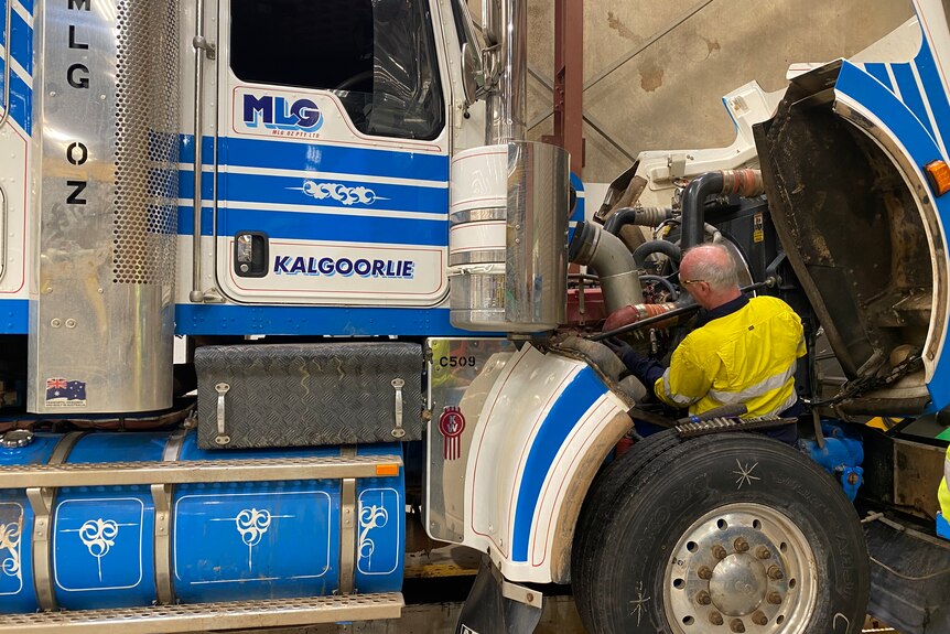 A man wearing a bright yellow shirt loos quite small as he works on repairing the the front of a large white and blue truck.