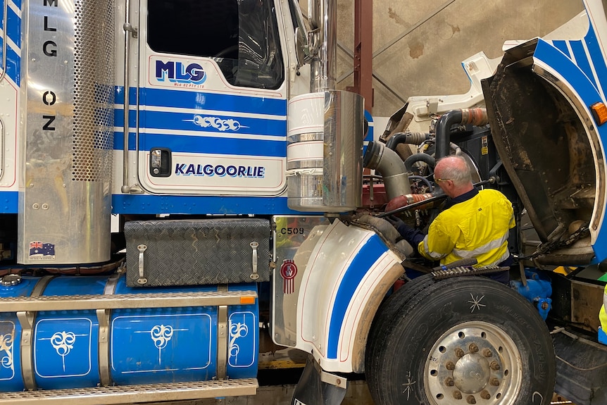 A man wearing a bright yellow shirt loos quite small as he works on repairing the front of a large white and blue truck.