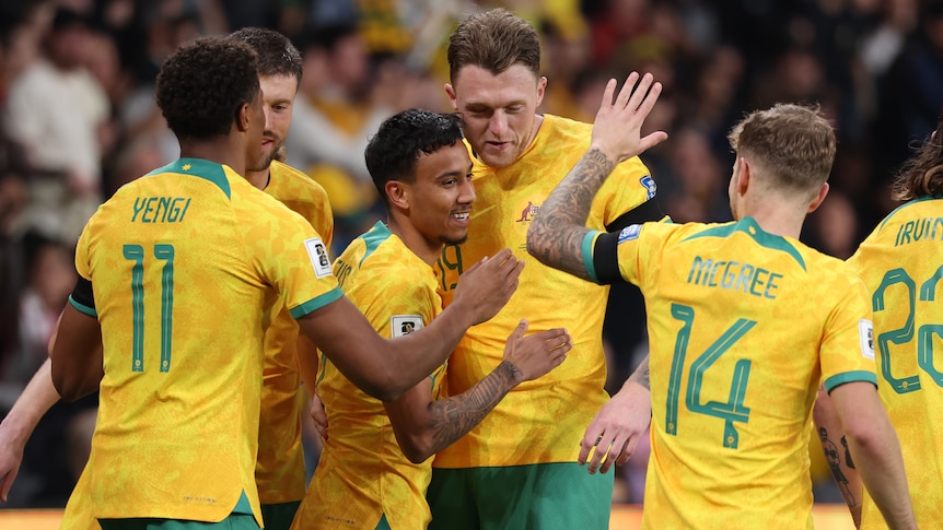 A Socceroos player is surrounded by teammates congratulating him after a goal.