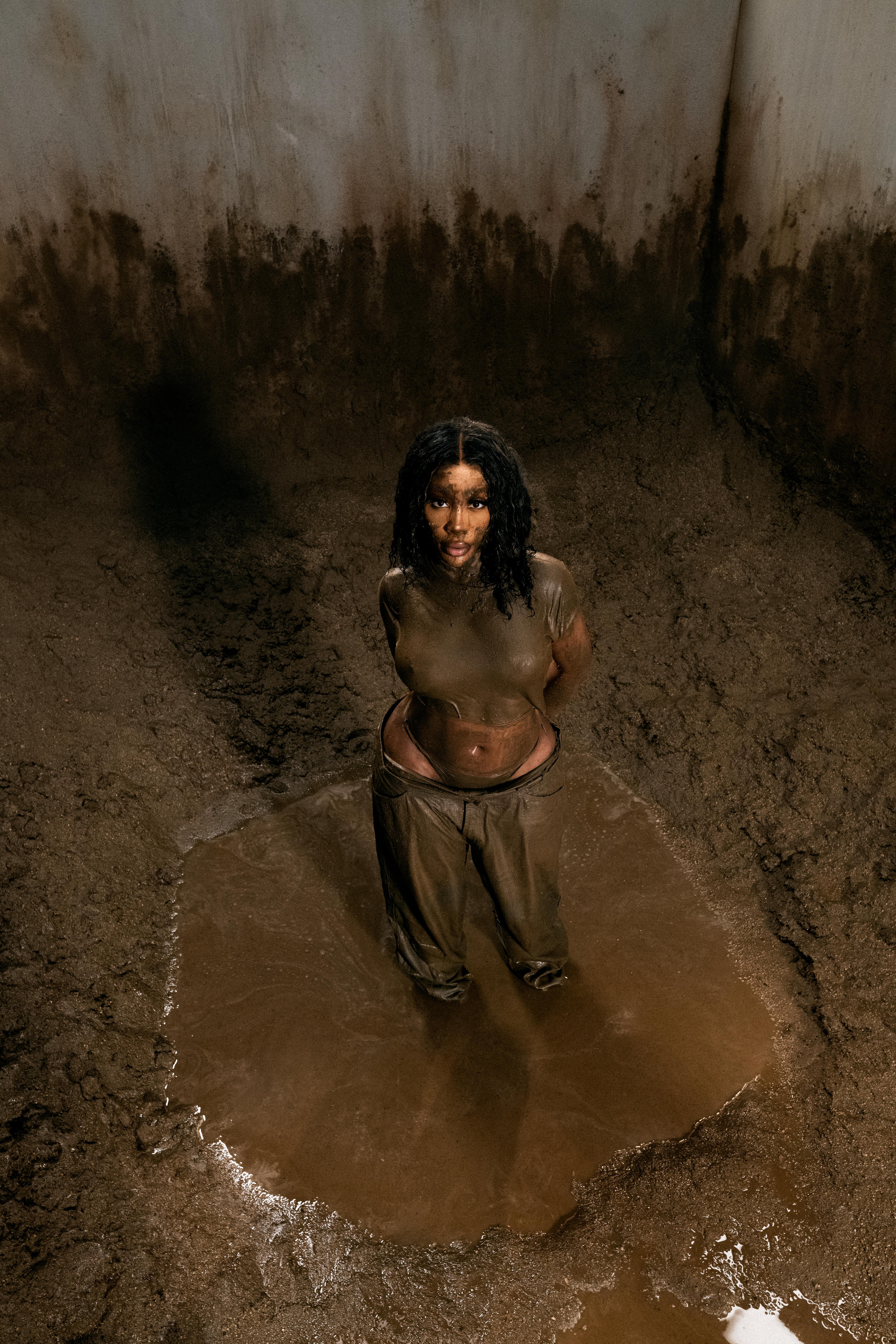 SZA is standing in the bottom of a muddy pit looking up with soaked clothes and dirt on her face
