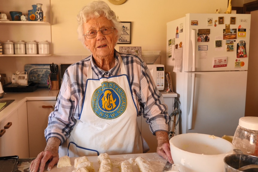 A lady stands in her kitchen, wearing a CWA apron, with raw scones in front of her, ready to put in the oven.