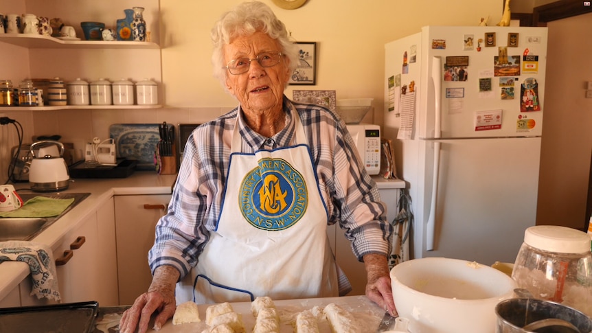 A lady stands in her kitchen, wearing a CWA apron, with raw scones in front of her, ready to put in the oven.