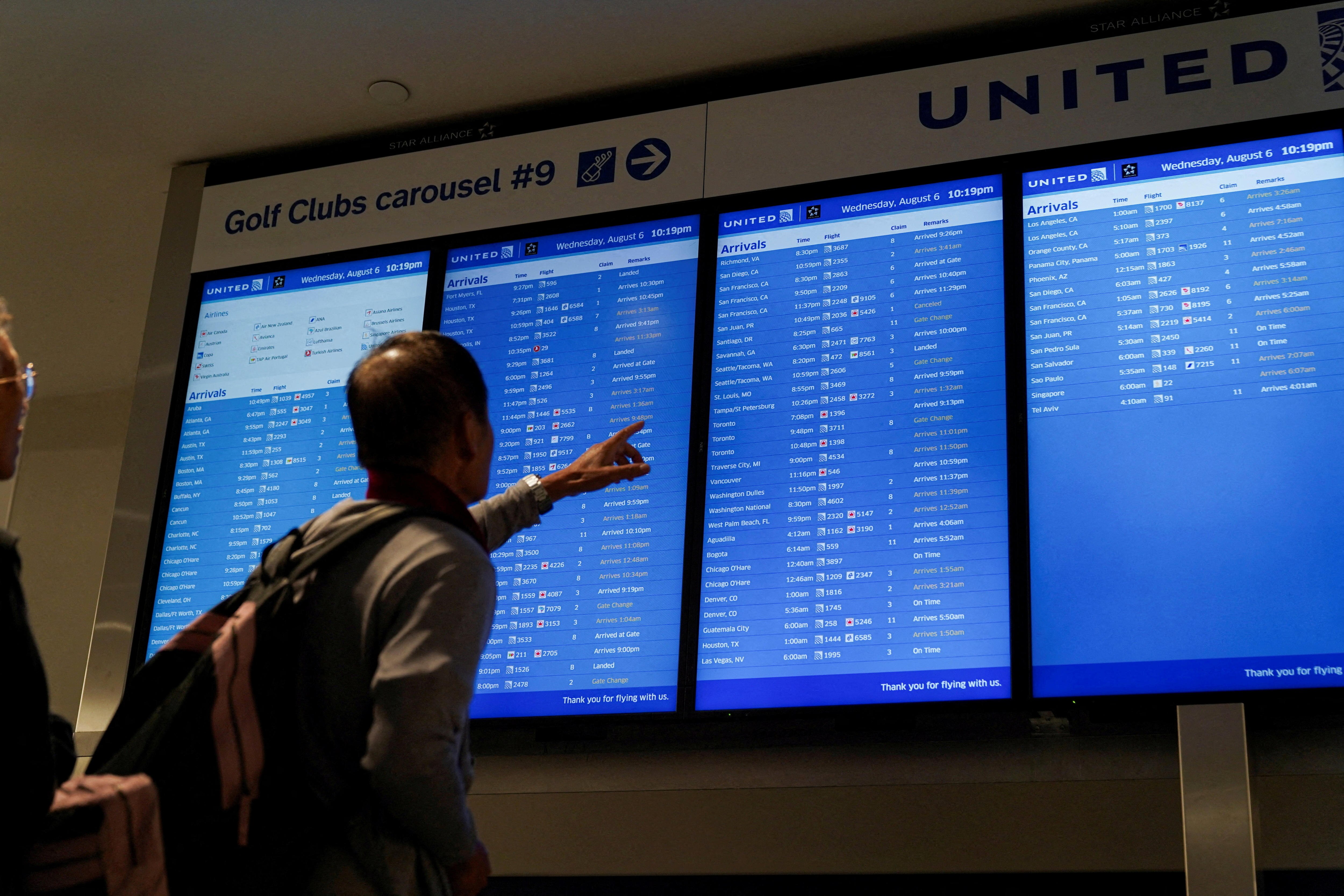 A man facing away with an arm and finger raised pointing at four blue flight arrival TV screens under a United banner