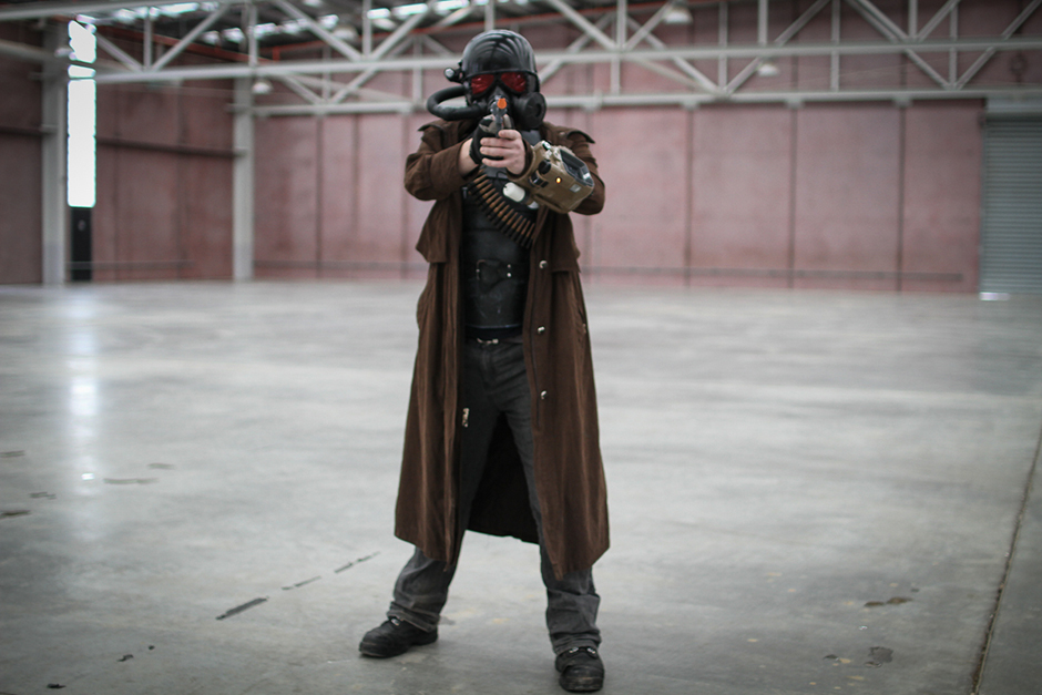 A man in costume, including a full-face black helmet and respirator, and long brown coat.