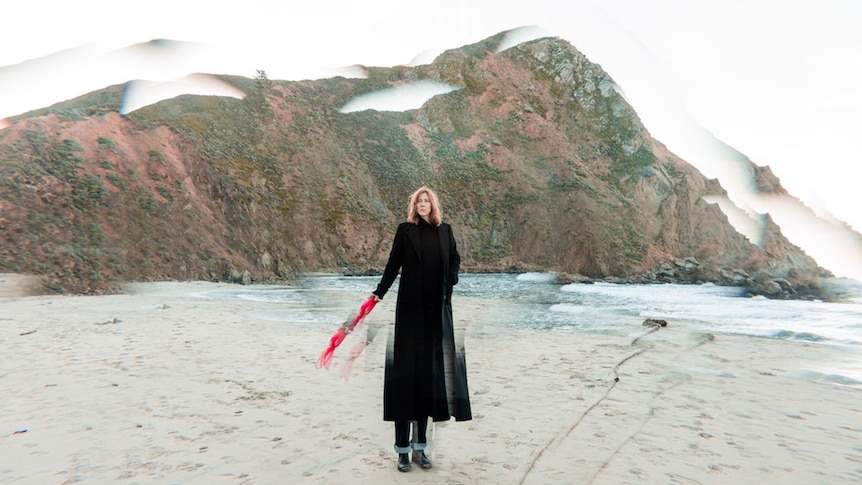 A woman is standing on a cold beach in a black dress with a small hill behind her. The image is distorted like broken glass.