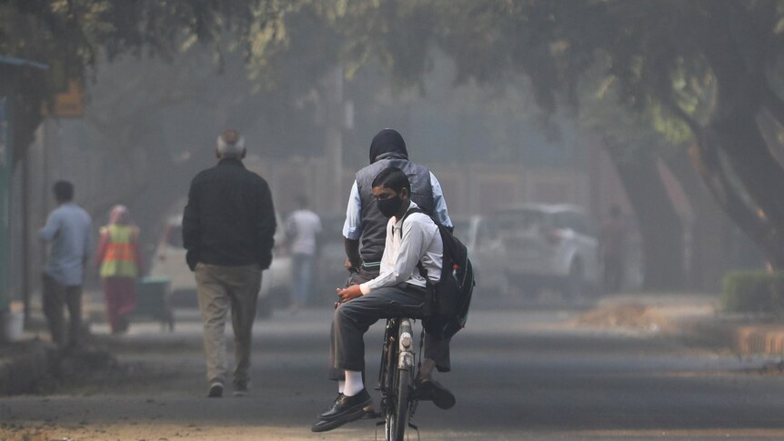 a boy in a suit shirt and pants, and a black mask, sits on the back of his father;s bike ona  smoggy road