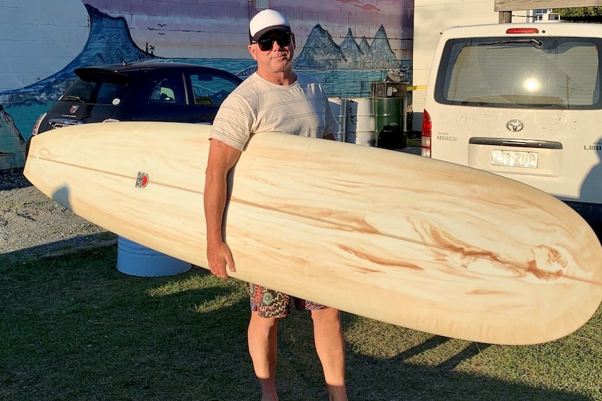 A middle-aged man in sunglasses and cap looks at camera while holding a long surfboard near beach