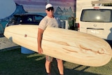 A middle-aged man in sunglasses and cap looks at camera while holding a long surfboard near beach