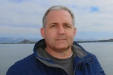 A man with grey hair dressed in a blue parka stands in front of a lake with mountains in the distance.