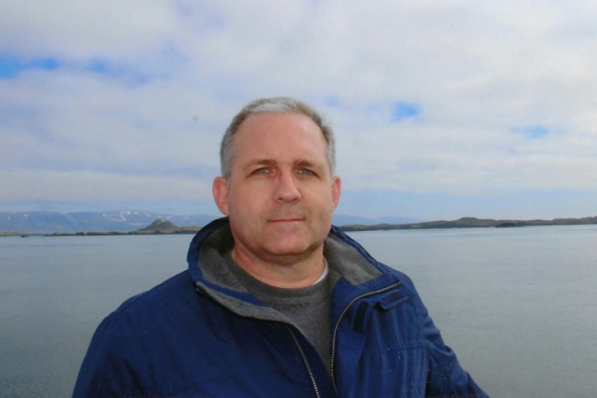 A man with grey hair dressed in a blue parka stands in front of a lake with mountains in the distance.