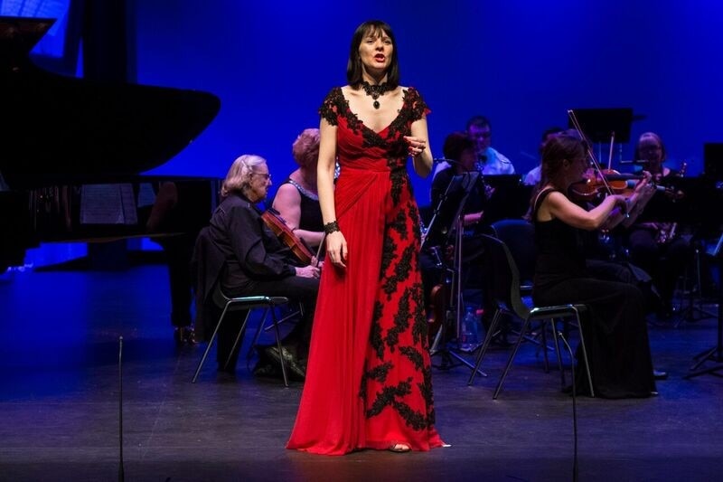 A woman in a long red dress sings with an orchestra behind her  