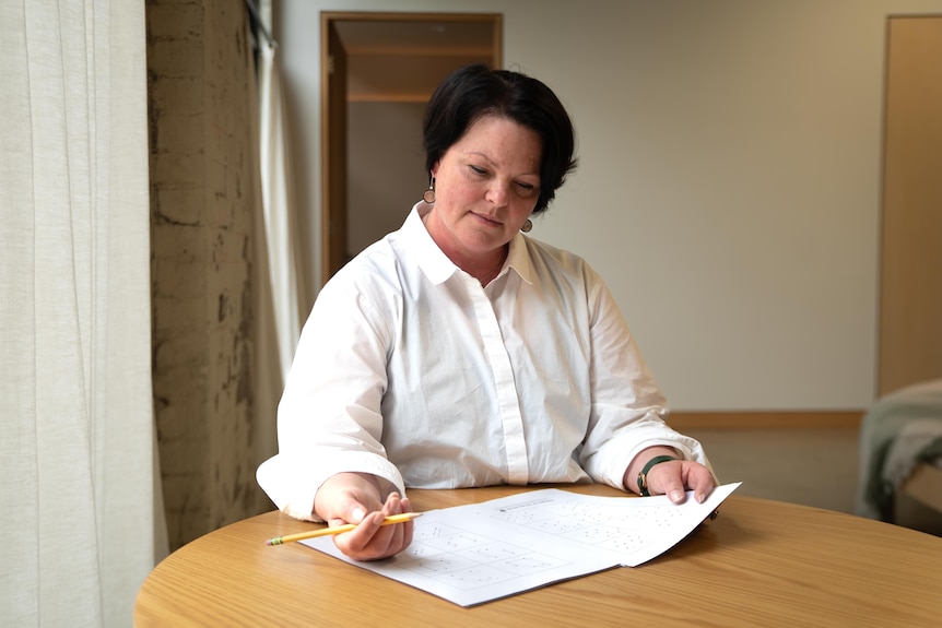 Natalie Foley, who has short brown hair and is wearing a white shirt, sits at a table with a pencil and several pages of paper