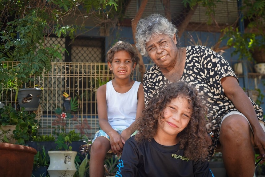 Grandma sits next to two grandsons in front of house 