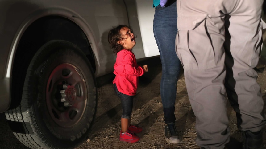 A child in a pink shirt cries and looks up at her mother, who is being detained by officials near the US-Mexico border.