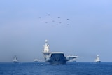 A aircraft carrier heads towards the camera. Smaller vessels flank it. In the sky above it are a number of jets.