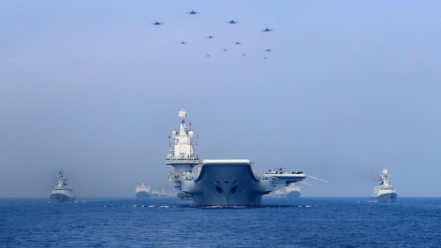 A aircraft carrier heads towards the camera. Smaller vessels flank it. In the sky above it are a number of jets.