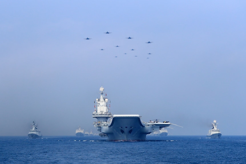 A aircraft carrier heads towards the camera. Smaller vessels flank it. In the sky above it are a number of jets.