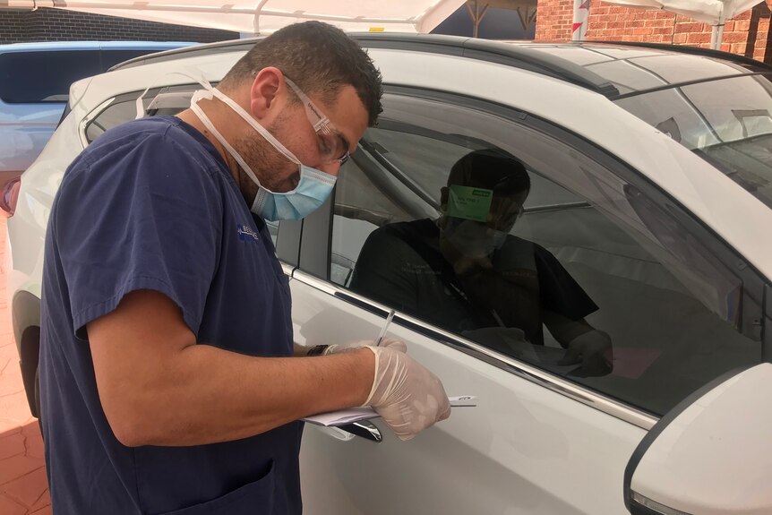 A man wearing scrubs, goggles and medical mask talks with someone through a car window