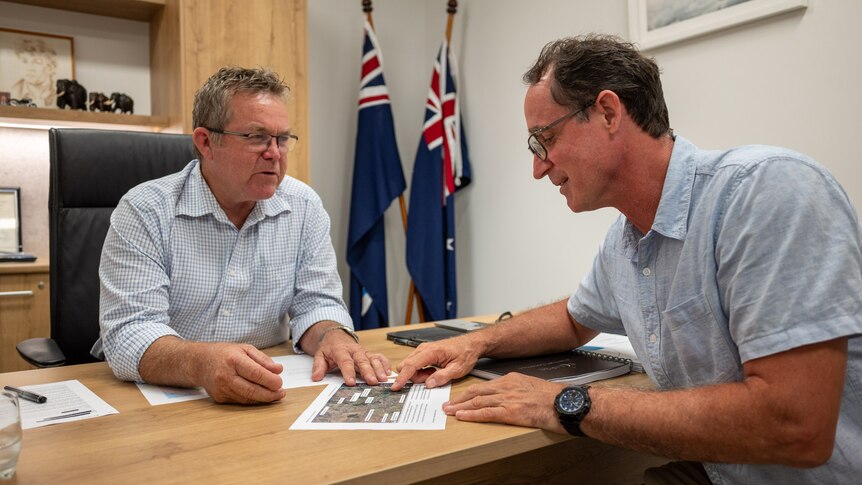 Two men sit at a desk talking and looking at pages in front of them. Between them in the background are two Australian flags.