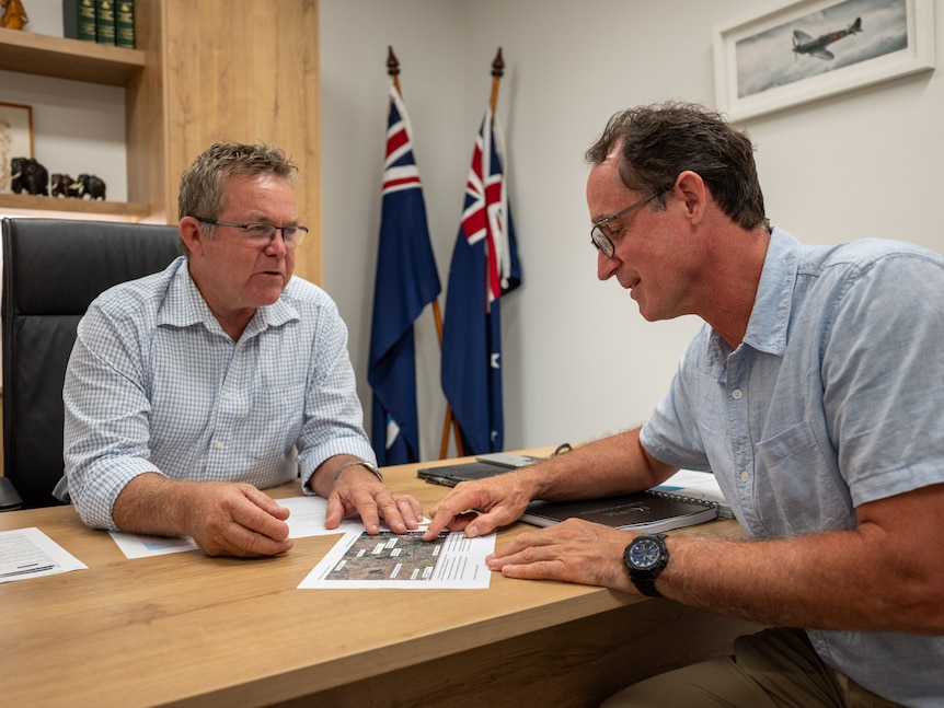 Two men sit at a desk talking and looking at pages in front of them. Between them in the background are two Australian flags.