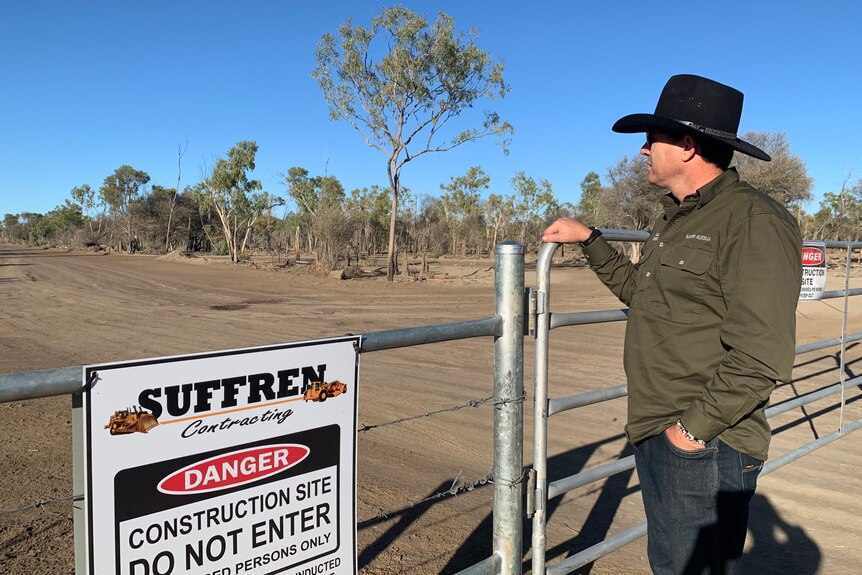 A man in a broad-brimmed hat leaning on the fence of a rural property and looking out into the distance.