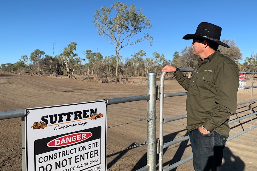 A man in a broad-brimmed hat leaning on the fence of a rural property and looking out into the distance.