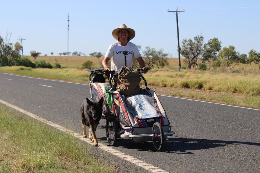 A man walking with two pram carriages and his pet dog on the side of a road