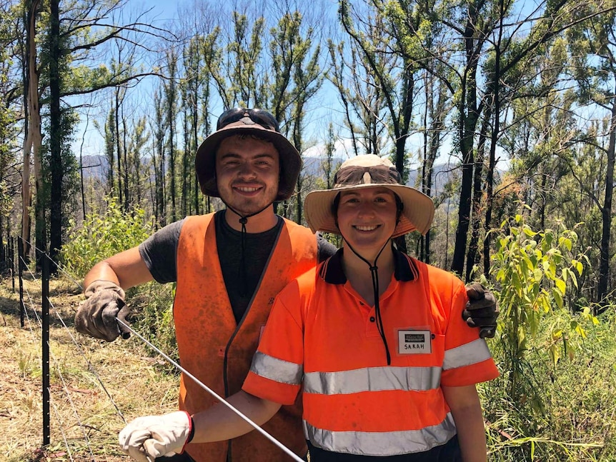 Two people in work clothes and hats standing in front of a fence.