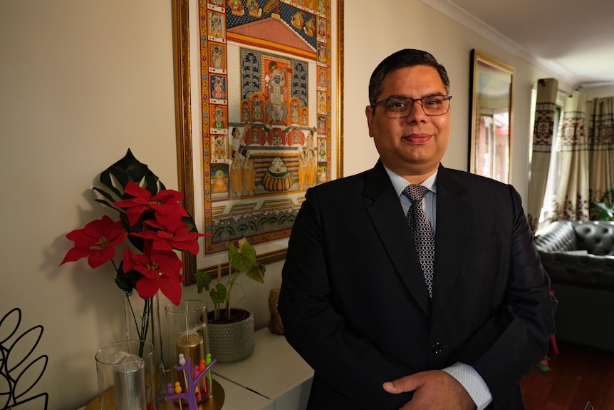 Mr Soni stands in his house wearing a suit in front of a colourful piece of art. 