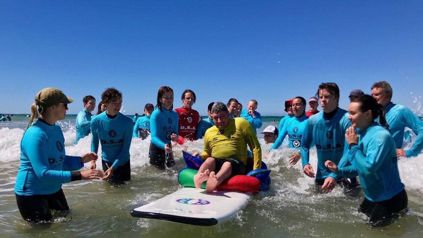 A young sits on a surfboard in the surf, surrounded by a team of volunteers.
