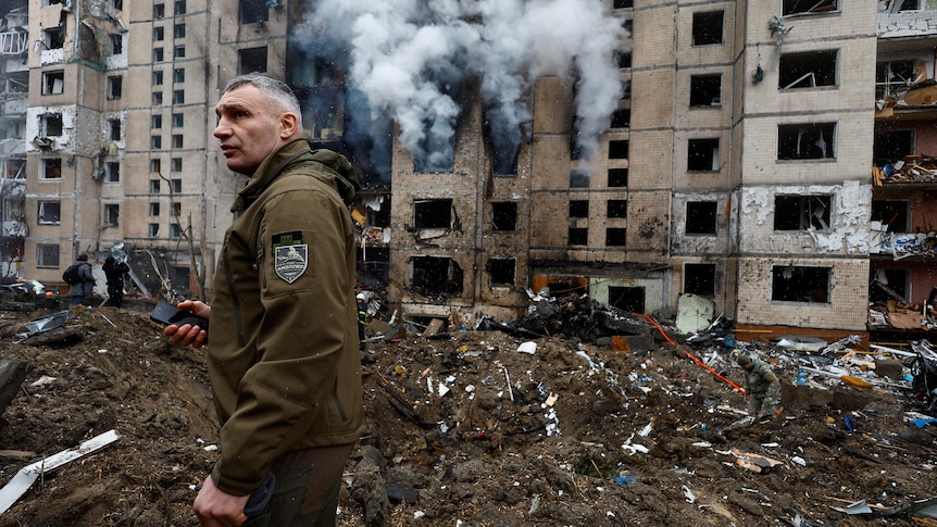 Man with army green jacket walks amid building smoke and debris 