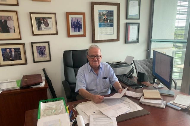 Sir Leo Hielscher in his home office in his retirement unit, sitting behind a desk, photos on the wall behind him.