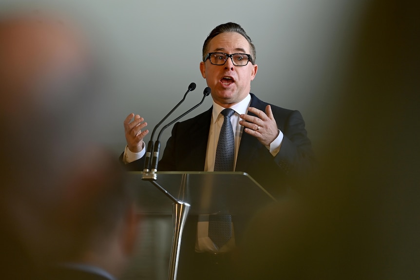 A man gestures widely with both hands while talking behind a lectern