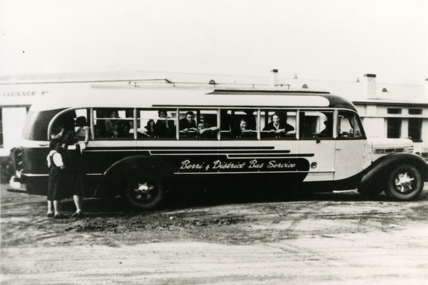A black and white image of an old 1950s style school bus, parked out the front of a school building.