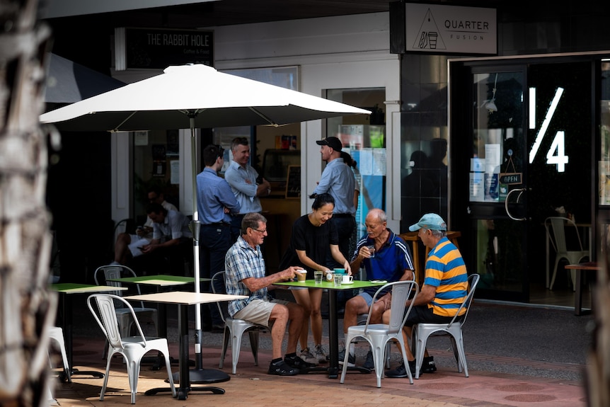  Outside a cafe, a waitress brings coffee to a group of men sitting at a table. No one is wearing a mask