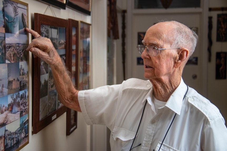 An elderly man looking at a wall full of framed photographs, in glasses and a white collared shirt