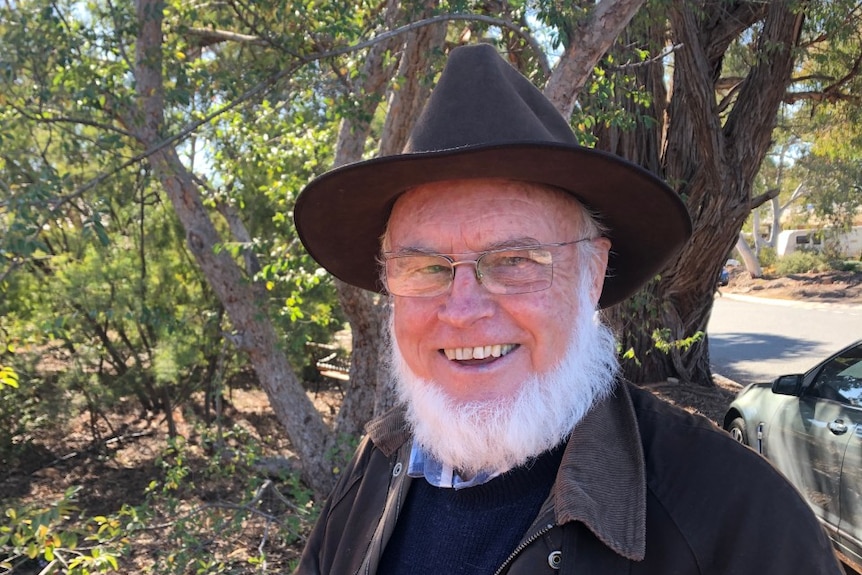 A man with a white beard and glasses smiles at the camera, with a tree behind him.